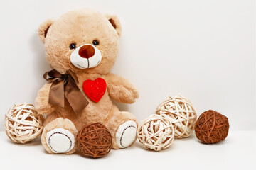 Cute teddy bear with a red heart on its chest sits near some wicker balls on a white background