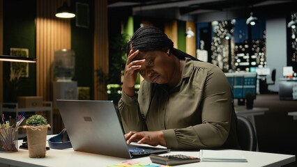 Black business employee realizing the night time alone in office, pushing limits despite clear signs of fatigue and work tension. Workplace challenges and sleep deprivation. Camera A.