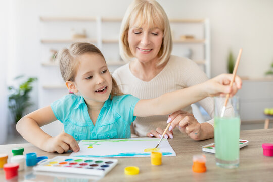 Elderly babysitter teaching little girl drawing, helping improve creative abilities, develop artist talent. Caring old 50s grandma spending time with small granddaughter at modern home