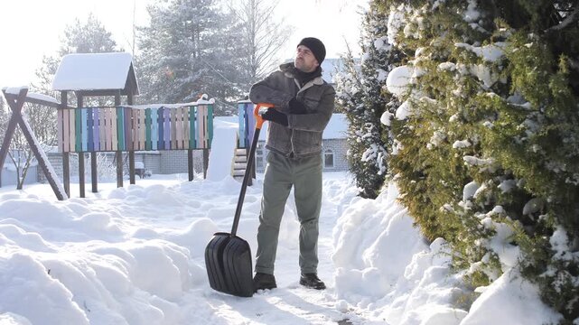 Man pausing on a sunny winter day while shoveling deep snow from a backyard path, standing with shovel, visible breath vapor, trees and house in bright cold light