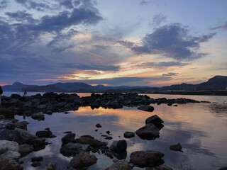 Fototapeta premium The image shows a calm pond with rocks in the foreground. Mountains are visible in the distance. The sky is colored in shades of sunset or dawn, with clouds.