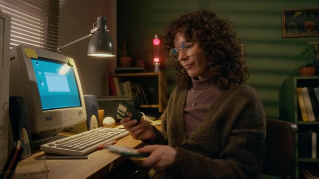 Medium shot of busy Caucasian woman in glasses, with curly hair searching stack of floppy disks, writing on label with necessary files, sitting at work desk at home with retro personal computer
