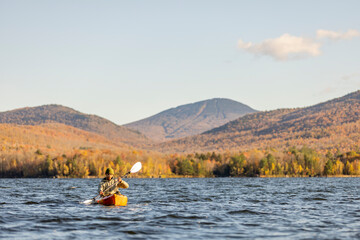 African-American man kayaking on a lake during peak fall autumn foliage with mountains and forest and view towards a ski area. Chittenden Reservoir, Vermont, New England, USA.