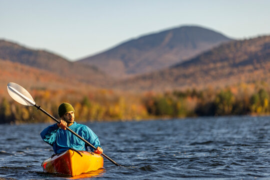 African-American man kayaking on a lake during peak fall autumn foliage with mountains and forest and view towards a ski area. Chittenden Reservoir, Vermont, New England, USA.