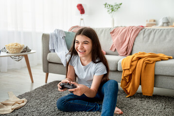 Happy Indian teenager with joystick playing online computer games in messy room during covid lockdown. Cheerful adolescent with controller engaged in video gaming at home © Prostock-studio