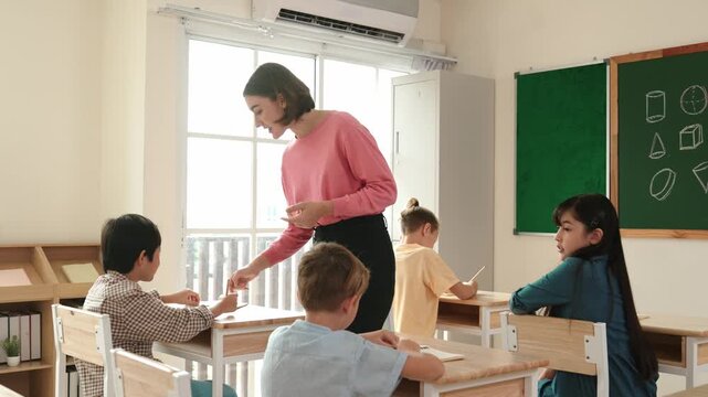 Caucasian teacher walking and check student homework at classroom while diverse children doing classwork at classroom. Diverse smart student lecturing and taking a note at elementary school. Pedagogy.