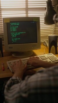 Vertical over-shoulder shot of male programmer in glasses and plaid shirt writing coding algorithm on vintage PC desktop computer while working in home office