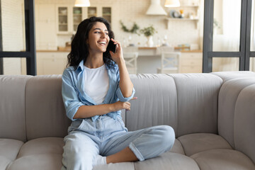 Obraz premium Happy young Caucasian lady sitting on couch, having mobile phone conversation at home, free space. Cheery millennial woman calling her friend or colleague on smartphone indoors