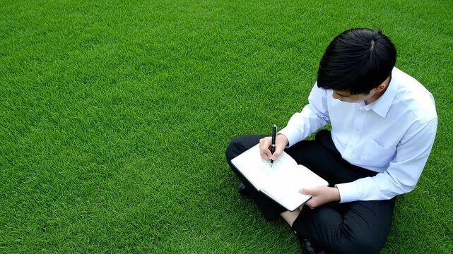 A male college student writing on a notebook while sitting in the grass in the university. University promotional video.