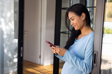 Mid-adult Asian woman leaning on sliding-door at home in blue top, holding maroon phone, copy space