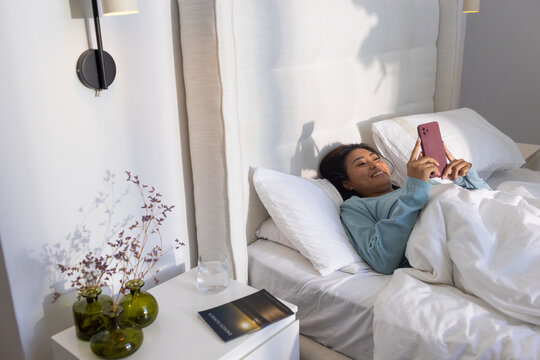 Mid-adult Asian female reclining on bed in lit bedroom, wearing blue top, holding pink smartphone