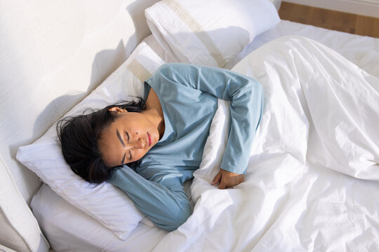 Mid adult Asian woman resting diagonally on bed under white duvet wearing light blue top