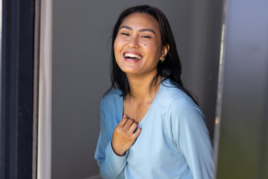 Mid adult Asian woman laughing, leaning in home doorway wearing light blue top and gold earrings