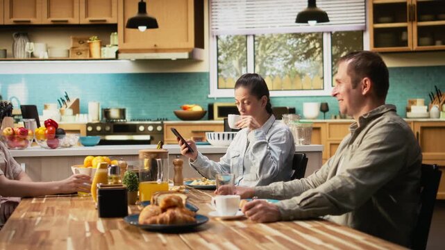 Preoccupied mother being detached and ignoring her family at the table, being distracted with a phone dependency and obsession during breakfast. Social media addiction. Camera A.