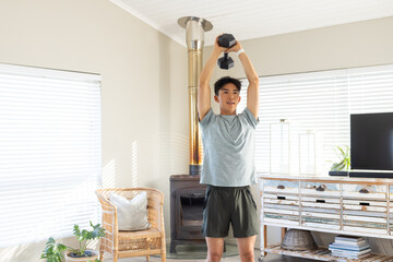 Asian man lifting hex dumbbell overhead in sportswear with fitness watch by living room wood stove