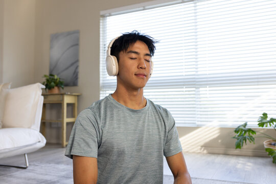 Asian man meditating on floor in living room, wearing white headphones and heather gray tee
