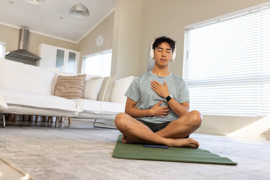 Asian man meditating on green yoga mat in living room, wearing headphones, smartphone on mat
