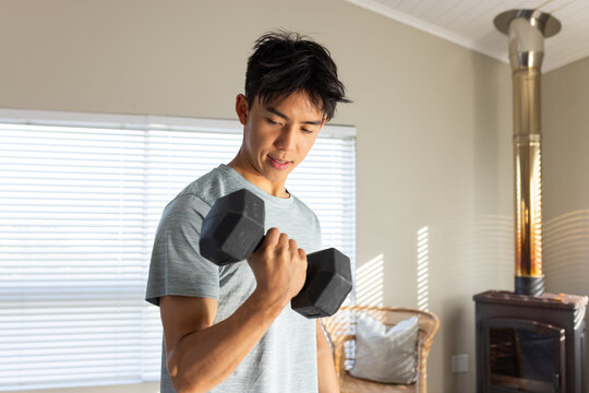 Man lifting black hex dumbbell and curling right arm in living room with blinds casting stripes