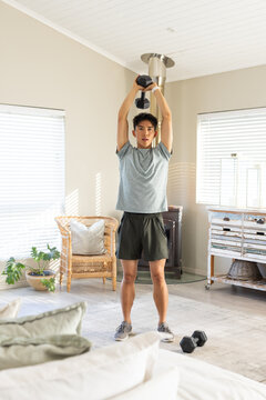 Adult Asian man lifting hex dumbbell overhead in living room in sportswear with second dumbbell