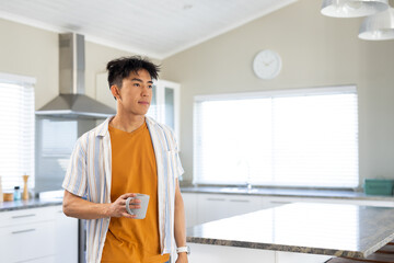 Adult Asian man standing by marble kitchen island, holding ceramic mug and gazing right
