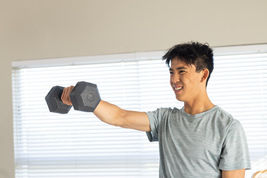 Asian man standing holding black hex dumbbell wearing light grey tee by closed blinds, copy space