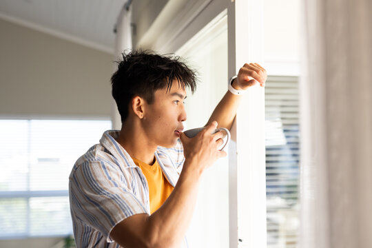 Asian man in his 20s standing at home by window holding white ceramic mug looking outside