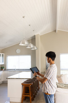 Asian man is using tablet in open-plan kitchen, smartwatch visible, island with pendant lights