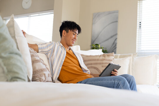 Asian man reclining on light sectional sofa, holding tablet, leaning on pillows near blinds