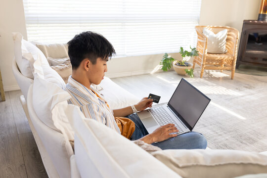 Asian man is entering card details while using silver laptop on white sectional couch near blinds