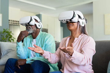 Senior couple wearing white VR headsets sitting on sofa in living room interacting virtually