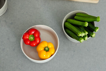 White bowls are sitting on gray countertop with red and yellow peppers, green zucchinis, wood board