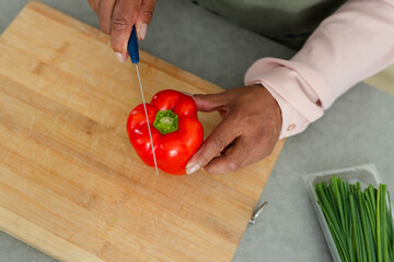 Red bell pepper is being sliced with blue-handled paring knife on gray countertop, chives nearby