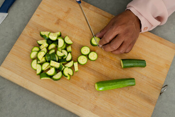 Zucchini is being sliced with chef's knife on wooden cutting board on gray countertop, pile left