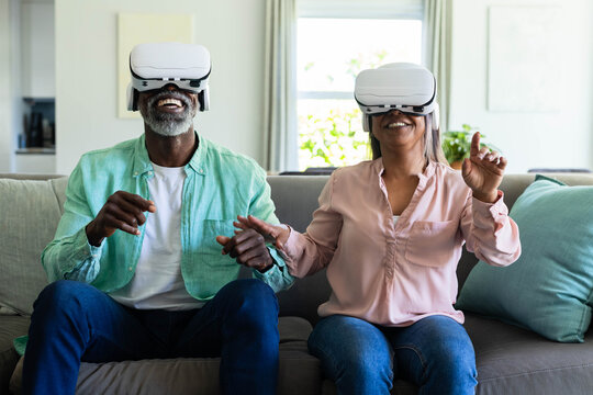 Diverse couple wearing white VR headsets, sitting and gesturing on gray couch in bright living room