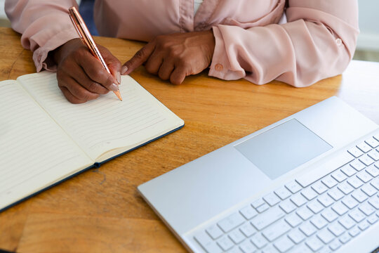 Metallic rose-gold pen is writing in lined notebook on wooden table, silver laptop nearby