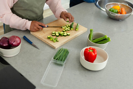 Hands and forearms are slicing zucchini on wooden board on gray kitchen countertop with bowls