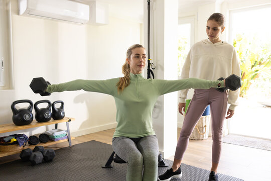 Female trainer and trainee performing lateral dumbbell raises on bench in home gym with kettlebells