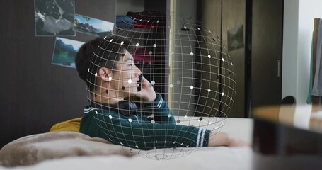 Reclining teen in green sweater holding phone to ear with headset on bed, AR globe overlay