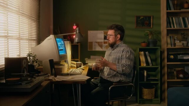 Medium full side shot of middle-aged male programmer sitting at work desk at home, trying to establish internet connection on retro computer with landline modem, while playing with ball during wait