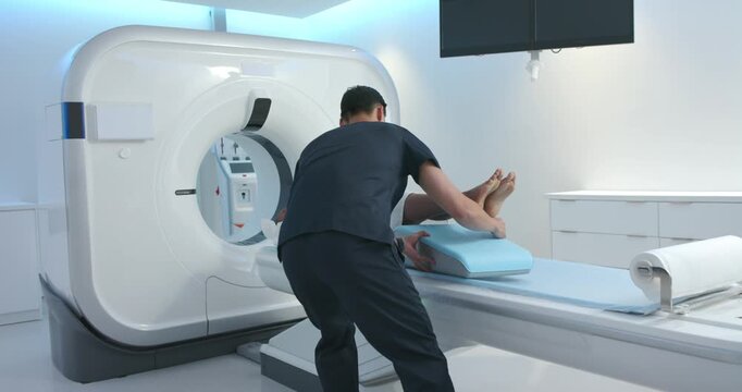 Technologist is grabbing light-blue leg wedge and is sliding it under patient's calves for CT scan