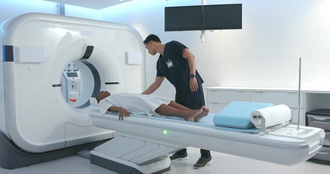 Female patient lying on scanner table, Asian tech placing blue wedge under calves, preparing scan