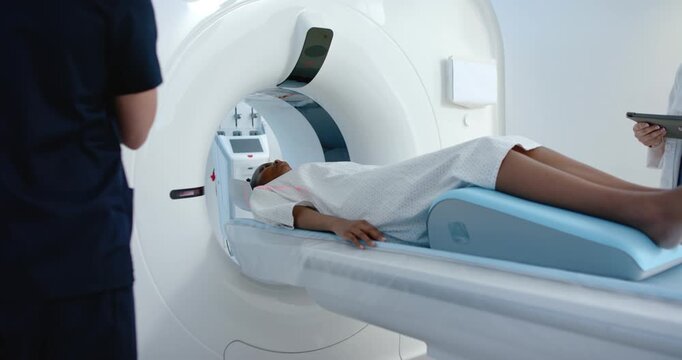 Patient in gown lying on exam table, sliding into scanner while tech operating panel starting scan
