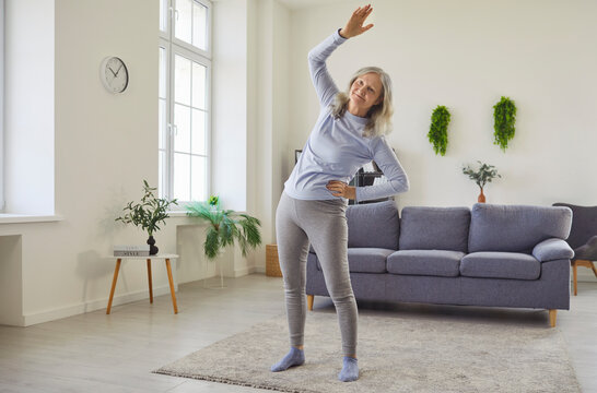 Happy senior woman doing physical exercise, side bends and stretching workout at home. Active old person training, grandmother standing on mat in living room with focus on mobility and balance posture