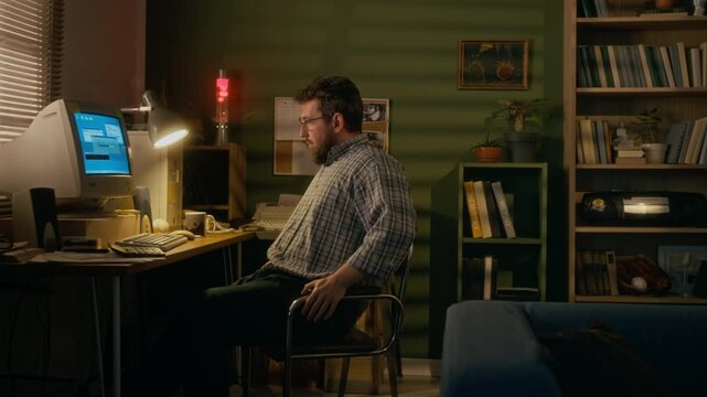 Full side shot of young Caucasian man sitting at desk at home, working on retro PC computer with CRT monitor, typing command and stretching in chair, while connecting to internet using landline modem