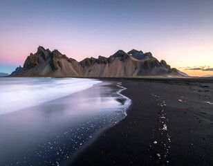 A scenic coastal view features a dark sand beach with long exposure water movement. Jagged mountains meet a colorful sky at dawn