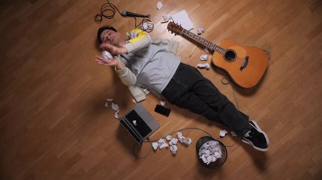 Musician lying on the floor surrounded by crumpled papers and music equipment, feeling frustrated and struggling with a lack of inspiration while trying to compose songs