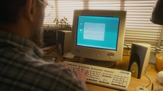 Over-shoulder shot of mature Caucasian man writing email on vintage desktop computer in home study, calculator, audio speakers, documents on table, communicating over internet, nostalgic 1990s vibe