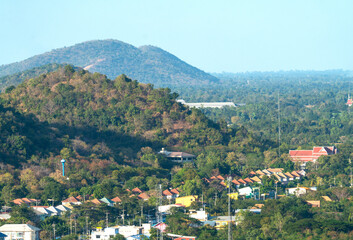 Scenic high angle view of a town in Thailand nestled among lush green tropical hills. Residential neighborhood with colorful roofs, local infrastructure, and dense forest under a clear blue sky.