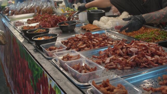 A display of popular Thai street food featuring chopped crispy pork belly (Moo Krob) and roasted red pork (Moo Daeng) in metal trays at a local market.