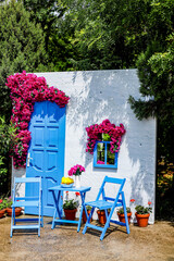 The photograph shows a dummy house with white walls, chairs and an entrance door with a blue window and beautiful red flowers hanging down.